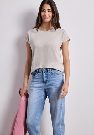 Kvinde iført lysegrå kortærmet top og blå jeans, der holder en sammenfoldet pink jakke i venstre hånd, stående mod en ensfarvet væg.