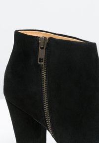 Botín negro de ante con detalle de cremallera metálica en el lateral, que presenta una textura suave y un diseño ajustado.