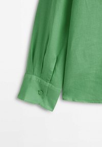 Manche de chemise en lin vert avec un poignet boutonné. Le tissu a une texture lisse avec des irrégularités subtiles. Le bouton est circulaire et cousu sur le poignet.