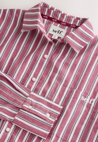 Camisa con botones a rayas verticales rojas, blancas y negras, con cuello, bolsillo delantero y marca "self:" en el bolsillo y etiqueta del cuello.