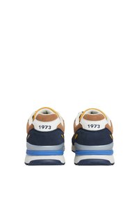 Sneaker con parte superiore in suede marrone e tessuto blu, un tallone bianco con la scritta "1973" e una suola in gomma blu. Elementi di design texturizzati visibili.