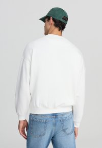 Felpa bianca in cotone con vestibilità comoda, polsini e orlo a costine. Abbinata a jeans in denim azzurri e un cappellino verde.