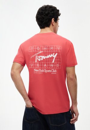 Man wearing coral red Tommy Jeans T-shirt with white grid and text "Tommy Jeans New York Sports Club" on back, paired with dark denim pants.