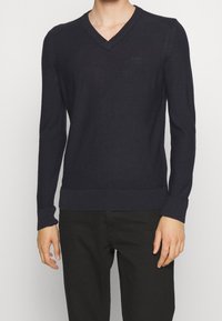 Navyblauer V-Ausschnitt-Pullover mit langen Ärmeln, gerippten Bündchen und Saum. Verfügt über ein dezentes Logo-Detail auf der Brust. Aus weichem, leichtem Material.