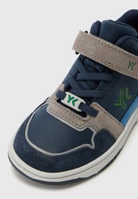 Sinine ja hall sportlik jalanõud, mille ülemine osa on valmistatud nubukist ja nahast, varustatud paeltega, Velcro rihmaga ja roheliste logo aktsentidega. Tekstuuriga tald.