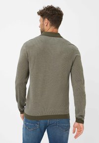 Olivgrüner, gestrickter Pullover mit geripptem Muster, langen Ärmeln und einem kontrastierenden, dunkleren grünen Kragen; kombiniert mit blauen Jeans.
