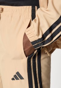 Pantalons de survêtement beige avec des rayures latérales noires, taille élastique et poche zippée. Fabriqués en tissu lisse et léger.