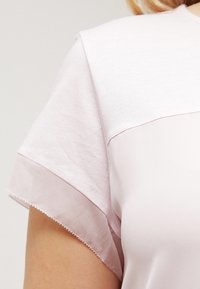 Lyserød top med korte ærmer, der har en tekstureret overdel og glat, gennemsigtig stof ved ærmerne, med en delikat bølget kant.