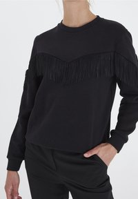 Černá mikina s texturovaným třásňovým vzorem přes prsa, kulatým výstřihem a dlouhými rukávy. Tkanina vypadá měkce a neformálně.