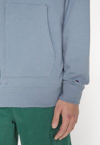 Světle modrý sweatshirt s přední kapsou, vroubkovanými manžetami a malým logem na rukávu, nošený přes texturované zelené kalhoty.