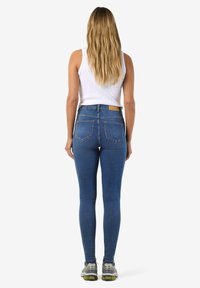 Femme aux longs cheveux blonds portant un haut sans manches blanc, un jean skinny bleu taille haute et des baskets gris-vert, debout de dos.