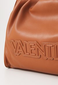 Rjava usnjena torbica z mehko teksturo, ki vsebuje izstopajoč embosiran logotip v drznih oblikah. Eleganten oblik s subtilnimi gubami.