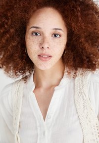White button-up shirt with a textured knit vest, featuring a round collar and an open neckline. Skin has freckles; dark, curly hair.