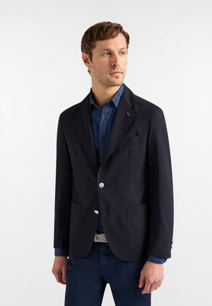 Mand iført en mørk marineblazer med sølvknapper, en blå mønstret skjorte og matchende mørke marinebukser, der kigger til siden.