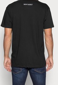 Svart t-shirt i bomull med korta ärmar, avslappnad passform och "NIGHT ADDICT"-logotyp i vitt på baksidan av halsen. Kombinerad med jeans.