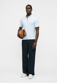 Homme en polo à manches courtes bleu clair, pantalon large et sombre, baskets blanches, tenant un sac en cuir beige plié devant un fond uni.