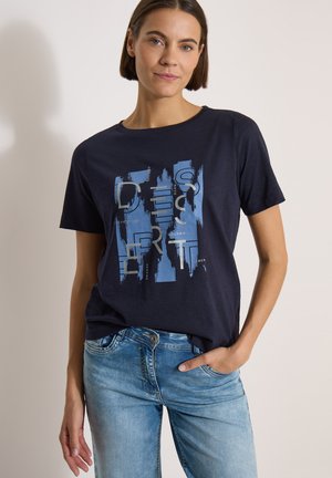 Femme aux cheveux bruns courts porte un t-shirt graphique bleu marine et un jean bleu clair, debout avec une main dans la poche devant un fond uni.