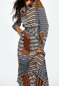 Femme portant une robe à manches longues avec un motif géométrique en orange, noir et blanc, accompagnée d'une ceinture assortie, posant avec une main sur la hanche.