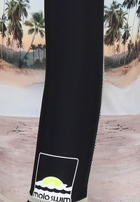 Mustat uimapuvut, joissa on kirkas trooppinen kuvio palmupuilla ja rannalla. Sisältää "molo swim" -logon alaosassa.