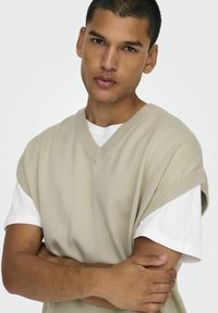 Strickweste in hellbeige mit V-Ausschnitt und weiten Armausschnitten, über einem weißen T-Shirt getragen. Der Stoff hat eine glatte Textur und gerippte Kanten.