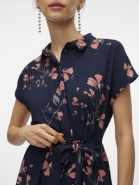 Žena koja podešava remen na struku tamnoplave haljine s uzorkom ružičastog cvijeća i kratkim rukavima, nosi viseće biserne naušnice.