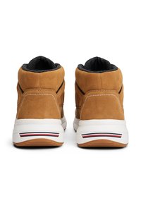 Une paire de baskets en daim beige avec des semelles blanches et des accents de rayures noires et rouges, vue de l'arrière sur fond blanc.