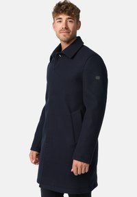 Marineblå uldfrakke med et klassisk design, bred krave, sidelommer og logoaccent på ærmet. Glat tekstur og struktureret silhuet.
