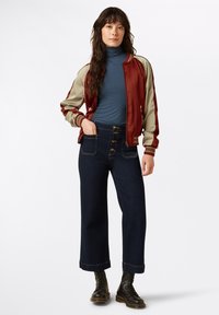 Red and beige satin bomber jacket over a blue ribbed turtleneck, paired with wide-leg dark denim jeans featuring button details and rolled cuffs.