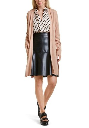 Femme portant un gilet beige ouvert, un chemisier à motifs, une jupe en cuir noire et des sandales compensées noires tressées, debout devant un fond blanc.