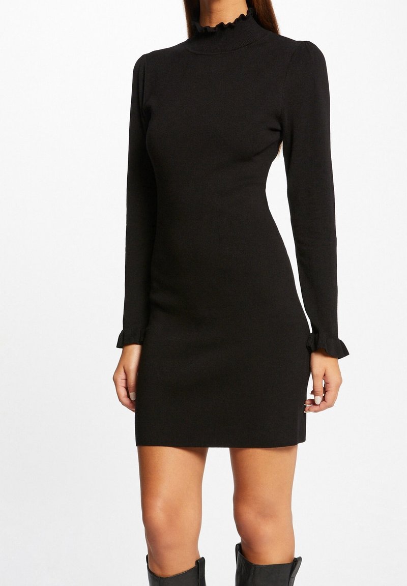 Robe noire à manches longues avec un col montant et des détails en volants, silhouette ajustée et tissu texturé. Ourlet au-dessus de mi-cuisse.