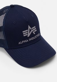 Tummansininen baseball-lippalakki, jossa on verkkopaneeli takana, ominaisuuksiin kuuluu litteä lippa ja keskeinen harmaa logo, jossa lukee "ALPHA INDUSTRIES" raidallisilla yksityiskohdilla.