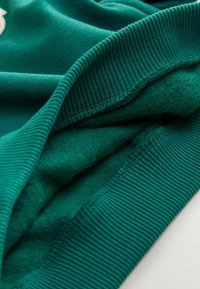 Gros plan sur une manche de vêtement en polaire verte, avec un poignet côtelé et des plis de tissu montrant la texture et les détails de couture.