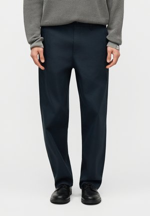 Άνδρας που φοράει σκούρο μπλε παντελόνι, γκρι πουλόβερ με υφή και μαύρα δερμάτινα επίσημα παπούτσια, στέκεται μπροστά σε απλό ανοιχτό φόντο.