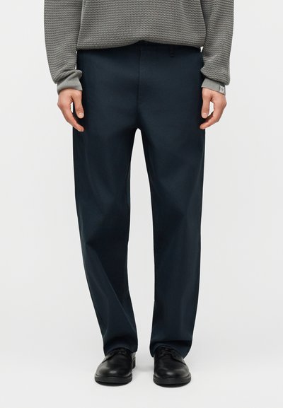 Homme portant un pantalon bleu marine foncé, un pull gris texturé et des chaussures habillées en cuir noir, debout devant un fond clair uni.
