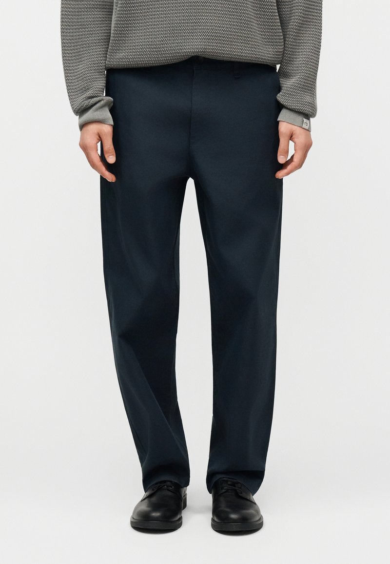 Άνδρας που φοράει σκούρο μπλε παντελόνι, γκρι πουλόβερ με υφή και μαύρα δερμάτινα επίσημα παπούτσια, στέκεται μπροστά σε απλό ανοιχτό φόντο.