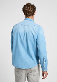 Camisa de denim de color azul claro con mangas largas, puños abotonados y cuello puntiagudo. Presenta una textura suave y un detalle de yugo en la parte posterior.