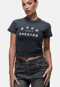 T-shirt gris coupé avec du texte blanc indiquant "GOLD + for NOTHING," col rond, manches courtes et tissu en coton lisse. Porté avec un jean bleu taille haute.