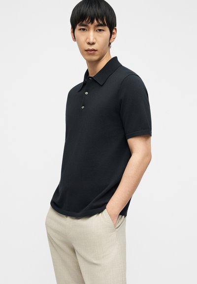 Νεαρός άνδρας με ίσια μαύρα μαλλιά, που φοράει μαύρο πόλο με κοντά μανίκια και μπεζ παντελόνι, στέκεται με το ένα χέρι στην τσέπη.