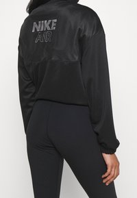 Bakifrån av en person i svart Nike Air-jacka och svarta leggings, stående mot en helt vit bakgrund.