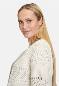 Gilet en tricot blanc avec des motifs texturés, un design en tissage ouvert et des manches larges, placé sur un fond neutre.