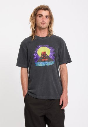 Jeune homme aux cheveux mi-longs portant un t-shirt noir avec un motif coloré de volcan sous une grande lune jaune.