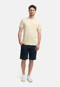 T-shirt beige à col ras du cou avec une poche sur la poitrine, associé à un short navy. Mannequin portant des baskets blanches ; design minimaliste, tissu léger.