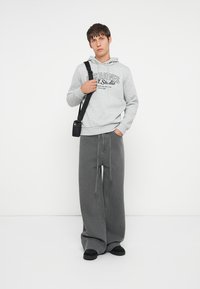 Sudadera gris con estampado de logotipo en el frente, combinada con pantalones grises de pierna ancha que presentan cordones. Un bolso cruzado negro y zapatos negros completan el conjunto.