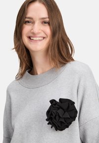 Grauer Pullover mit einem gerippten Ausschnitt und einem auffälligen schwarzen Stoffblütenakzent auf der linken Seite. Weiche Textur und lockere Passform.