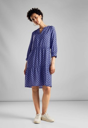 Blau gemustertes Kleid mit V-Ausschnitt, 3/4-Ärmeln, geraffter Taille und gestuftem Saum, kombiniert mit hellen Sneakers.