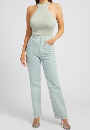 Vrouw draagt een mouwloos geribd haltertopje, lichtblauwe hooggetailleerde spijkerbroek met patroon en sandalen met hoge hakken en bandjes, staand met één hand in de zak.