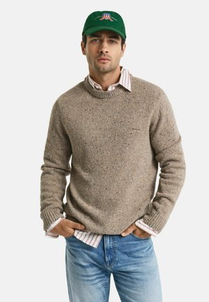 Mann mit grüner Mütze, beigem Strickpullover über einem gestreiften Hemd und blauen Jeans, mit den Händen in den Taschen, der frontal vor einem weißen Hintergrund steht.