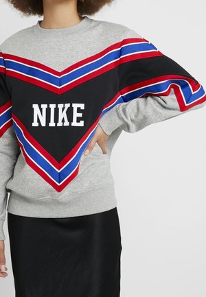 Osoba w szarej bluzie Nike z wyrazistymi czerwonymi, niebieskimi i białymi paskami w jodełkę, w połączeniu z czarną spódnicą, ręka na biodrze.