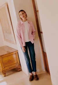 Tânără sprijinită de perete, purtând o cămașă cu dungi roz, top alb, blugi închiși la culoare și mocasini maro, într-o cameră iluminată difuz, cu mobilier din lemn.