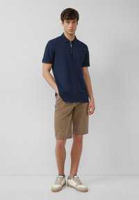 Granatowa koszulka polo z krótkim rękawem i zamkiem, w połączeniu z khaki szortami. Model nosi beżowe i białe sneakersy. Gładka tekstura materiału.